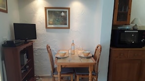 A dining area with a wooden table, four chairs, a painting on the wall, a television on a stand, and a microwave in the kitchen area.