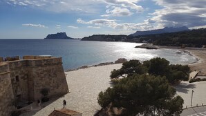 Udsigt over kysten med en bygning i sten, en strand og et bjerg i baggrunden.