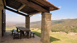 Un patio de piedra con una mesa y bancos de madera, con vistas a un valle y colinas lejanas bajo un cielo despejado.