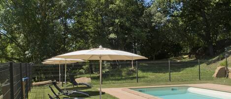 A poolside area with lounge chairs and umbrellas, surrounded by a fence and trees.