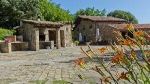 Une cour en pierre, avec des bâtiments à l’aspect rustique, un chemin pavé et des plantes en fleurs.