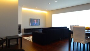 A modern living room with a black sofa, a flat-screen TV mounted on the wall, a dining table with chairs, and a fruit bowl on the table. - Affordable Luxury Beach Apartment with superb sea view 10 minutes from Porto (Vila Nova de Gaia)