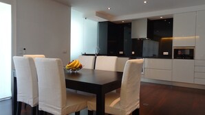 A modern dining area with a dark wooden table and white upholstered chairs. A fruit bowl with bananas and apples is on the table. The room has a minimalist design with white walls and dark cabinetry. - Affordable Luxury Beach Apartment with superb sea view 10 minutes from Porto (Vila Nova de Gaia)