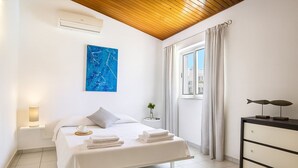 Una habitación de hotel moderna con una cama, una mesita de noche con lámpara, una ventana con cortinas, un aire acondicionado instalado en la pared y un cuadro abstracto azul.