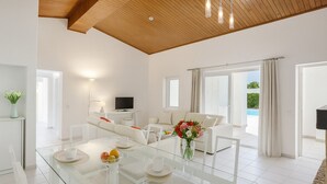 Una sala de estar moderna con una mesa de comedor de vidrio, sofás blancos, un televisor de pantalla plana y vista a una piscina.