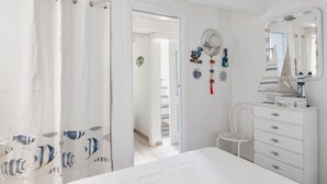 A white bedroom with a bed, a dresser, a chair, and a mirror. There is a door leading to another room. - To complete (Porto Rotondo)