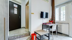 Una stanza con una porta scura, una TV a muro, un tavolino con una sedia e una finestra con tende.
