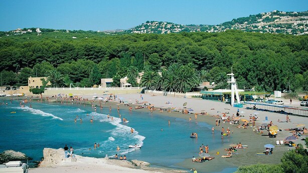 Une plage où l’on voit des gens nager et bronzer, une tour de surveillance des sauveteurs, et une colline avec des bâtiments en arrière-plan.