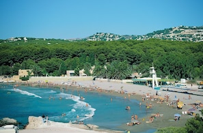 Ein Strand mit badenden und sonnenden Personen, einem Rettungsturm und einer Hügelkette mit Gebäuden im Hintergrund.