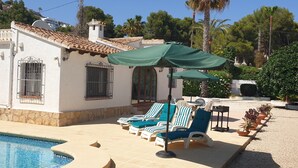 Un espace aménagé au bord de la piscine, avec des chaises longues et un parasol vert, un bâtiment blanc doté d’un toit en tuiles et un ciel bleu azur.