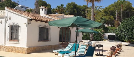 A poolside area with lounge chairs and a green umbrella, a white building with a tiled roof, and a clear blue sky.