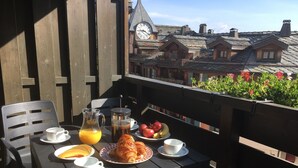 Una mesa de madera con alimentos para el desayuno, como croissants, fruta y café, en un balcón con vistas a edificios tradicionales.