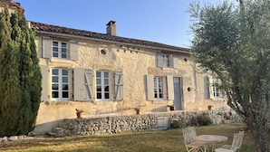 Une maison de campagne en pierre, dotée de nombreuses fenêtres et volets, d’un mur en pierre et d’un petit espace extérieur aménagé avec une table et des chaises.