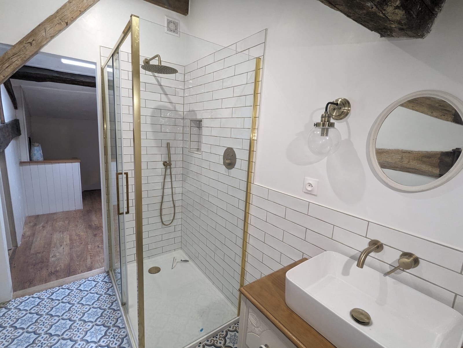 A modern bathroom with a glass-enclosed shower, a white sink with gold fixtures, and a round mirror.