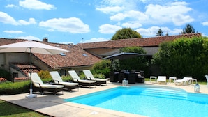 Une piscine entourée de chaises longues et de parasols, elle-même bordée d’un jardin et d’un bâtiment dont le toit est recouvert de tuiles.