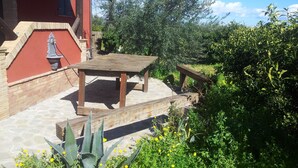 Un patio con una mesa y sillas de madera, rodeado de follaje y un edificio rojo.