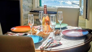 Mesa lista para una comida con botella de vino, copas, platos y cubiertos. Se ve una ventana con vistas al océano al fondo.