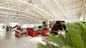 Ampla sala de estar com pé direito alto, sofás vermelhos, um piano de cauda e uma grande janela que proporciona iluminação natural.
