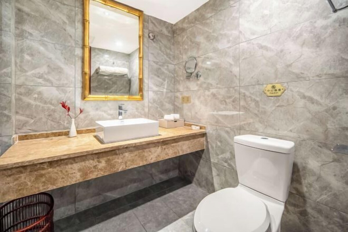 A modern bathroom with a marble wall, a wooden vanity, a white sink, a toilet, and a mirror with a gold frame.