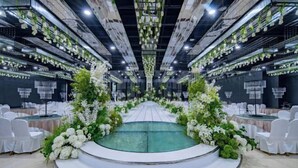 Un espace doté d’une scène centrale, entouré de chaises et de tables blanches, agrémenté de compositions florales vertes et de guirlandes lumineuses suspendues.