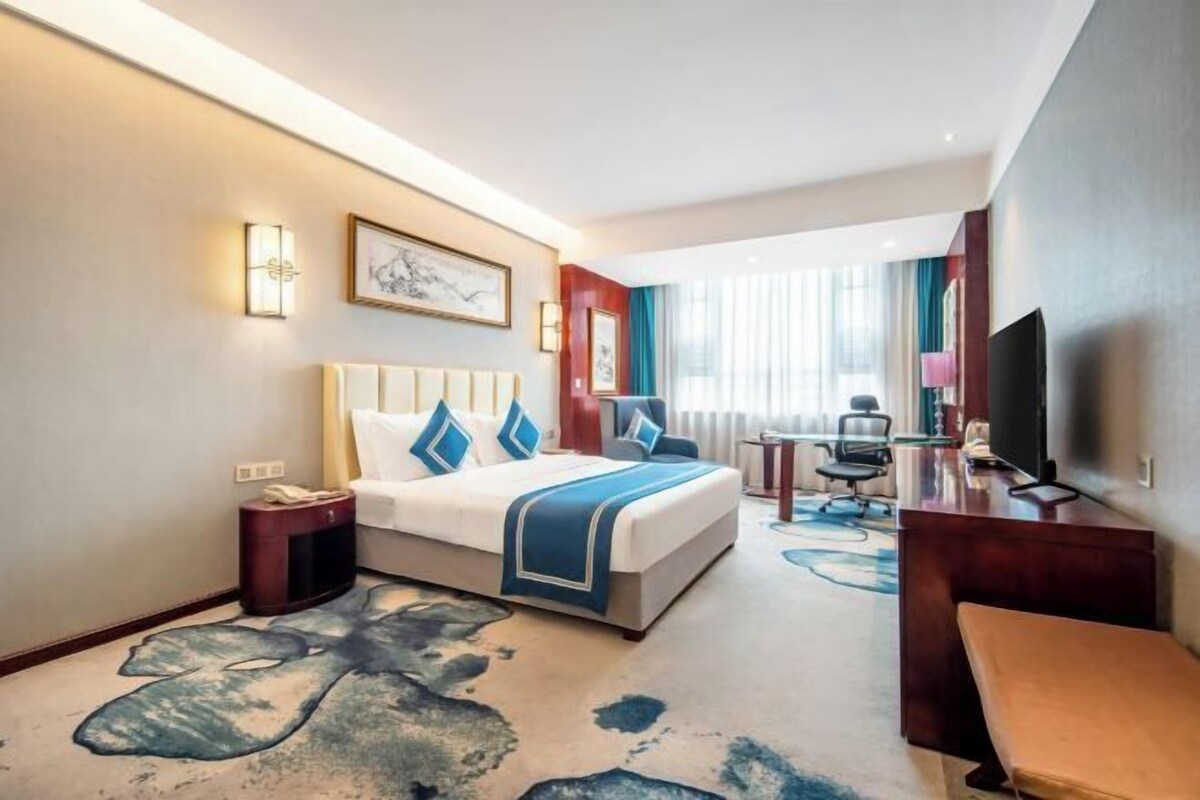 A hotel room with a bed, bedside tables, a desk, a chair, a television, and a large window with curtains.