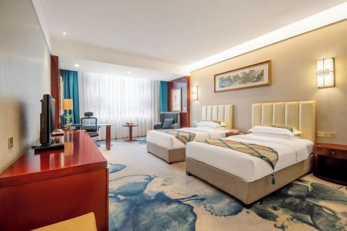 A hotel room with two beds, a desk, a TV, and a large window with curtains.