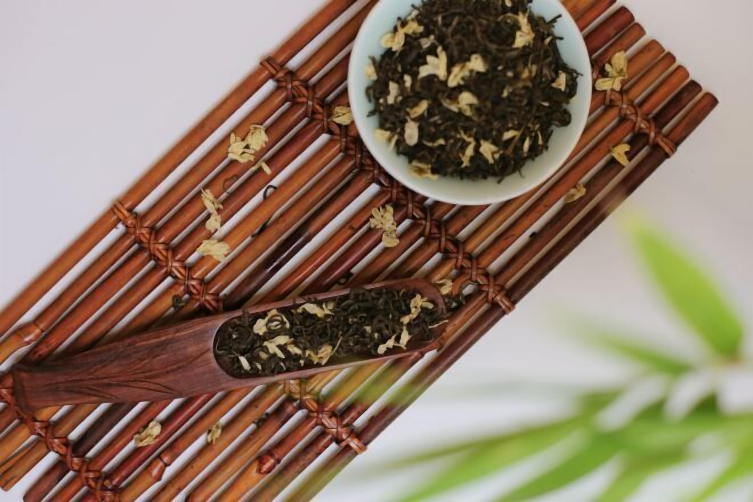 A bamboo mat with a wooden spoon containing tea leaves and a bowl of tea leaves.