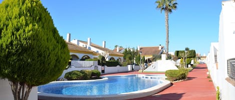 A swimming pool with a red pathway, surrounded by white buildings with tiled roofs and palm trees.