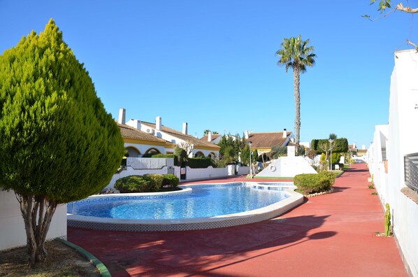 A swimming pool with a red pathway, surrounded by white buildings with tiled roofs and palm trees.