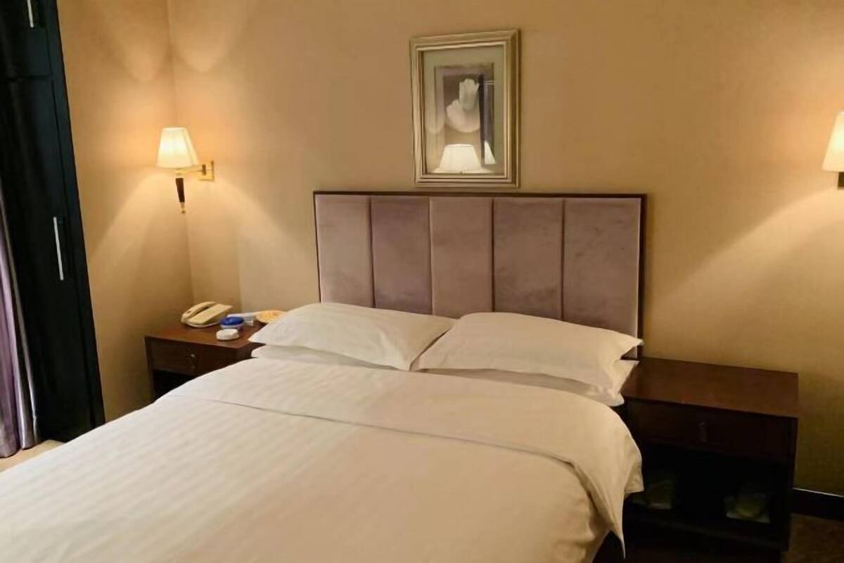 A hotel room with a bed, bedside lamps, a framed picture on the wall, and a nightstand with a phone and a lamp.