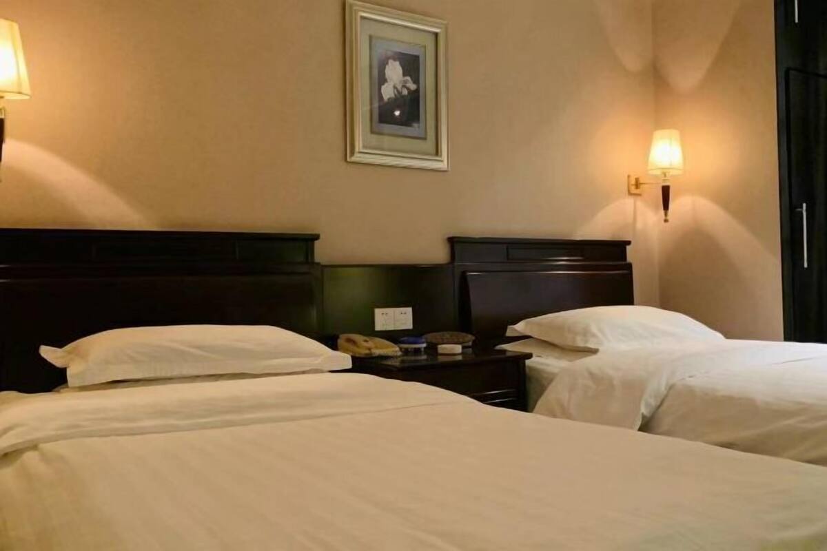 A hotel room with two single beds, a nightstand with a lamp, and a framed picture on the wall.
