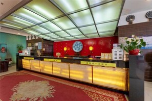 Zona de recepción de un hotel con techo de vidrio, pared roja con un plato decorativo y mostrador de recepción con una lámpara roja.