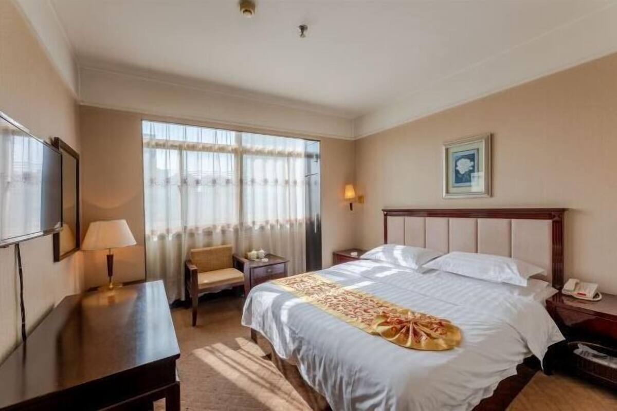A hotel room with a bed, a desk, a chair, a lamp, a bedside table, and a window with curtains.
