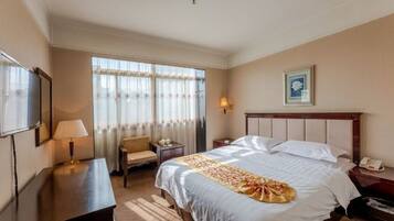 A hotel room with a bed, a desk, a chair, a lamp, a bedside table, and a window with curtains.