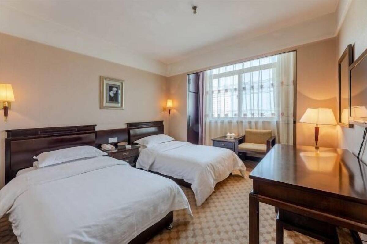 A hotel room with two beds, a desk, a chair, a lamp, a picture on the wall, and a window with curtains.