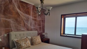 A bedroom with a bed, a nightstand, a lamp, a chandelier, and a window with a view of the sea. - Beachfront Apartment - By the SEA (Vila do Conde)