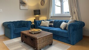 A living room with a blue sofa set, a wicker coffee table, and a large window with curtains. - Sunny, spacious & fully equipped, castle views, close to lake/beach, restaurants (Schwangau)