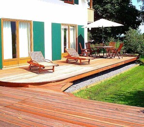 Une terrasse en bois, dotée de chaises longues et d’une table, jouxtant un bâtiment aux volets verts et à la façade blanche.