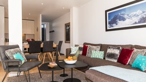 Un salon moderne avec un canapé marron, des fauteuils gris, une table basse et un coin repas comprenant une table et des chaises en bois.
