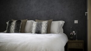 Un lit bien fait, avec du linge de lit blanc et des coussins à motifs, une petite table de chevet avec une lampe et une plante en pot, et un mur sombre à la texture marquée.