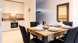 Une cuisine moderne avec des armoires blanches et un four encastré, un coin repas avec une table en bois prévue pour quatre personnes, et un grand miroir au mur.