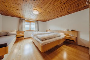 Een slaapkamer met een houten plafond, een bed met witte beddengoed, een nachtkastje, een raam met gordijnen, en een houten vloer.