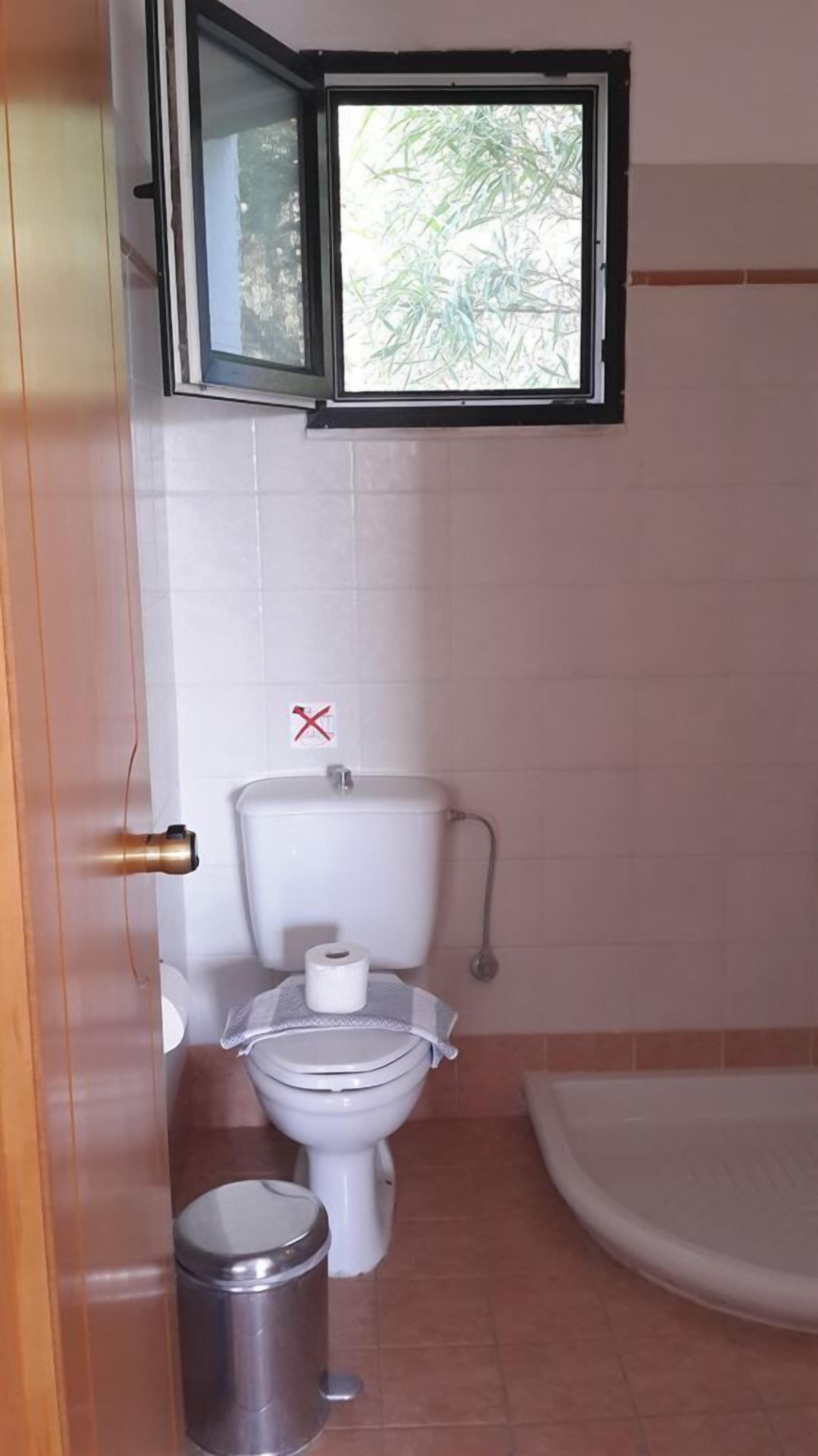 Une petite salle de bain avec des toilettes, une poubelle et une fenêtre située au-dessus des toilettes.