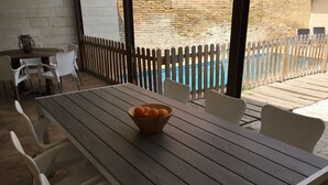 Una mesa de madera con un bol de naranjas, sillas blancas y vistas a una piscina a través de una valla de madera.