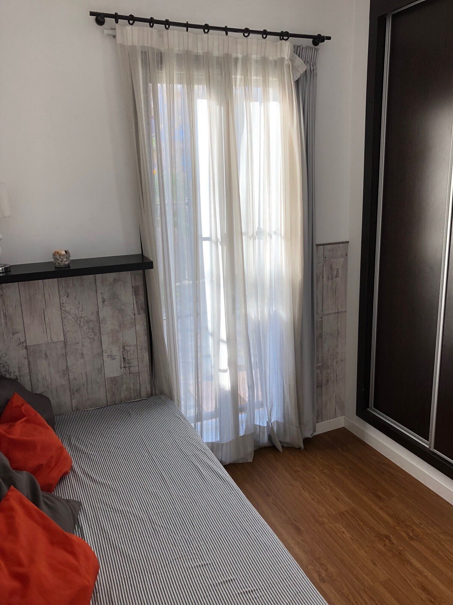 A room with a bed, a wooden floor, a black wardrobe, and sheer curtains.