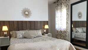 Una cama bien hecha con colcha y almohadones estampados, dos lámparas de noche, un espejo grande y una ventana con cortinas florales.