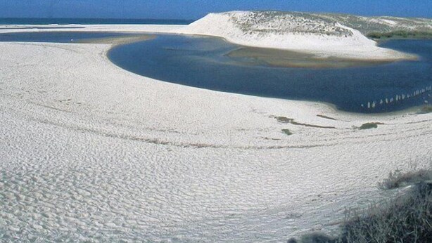 Un extenso paisaje arenoso con una gran masa de agua y una colina al fondo.
