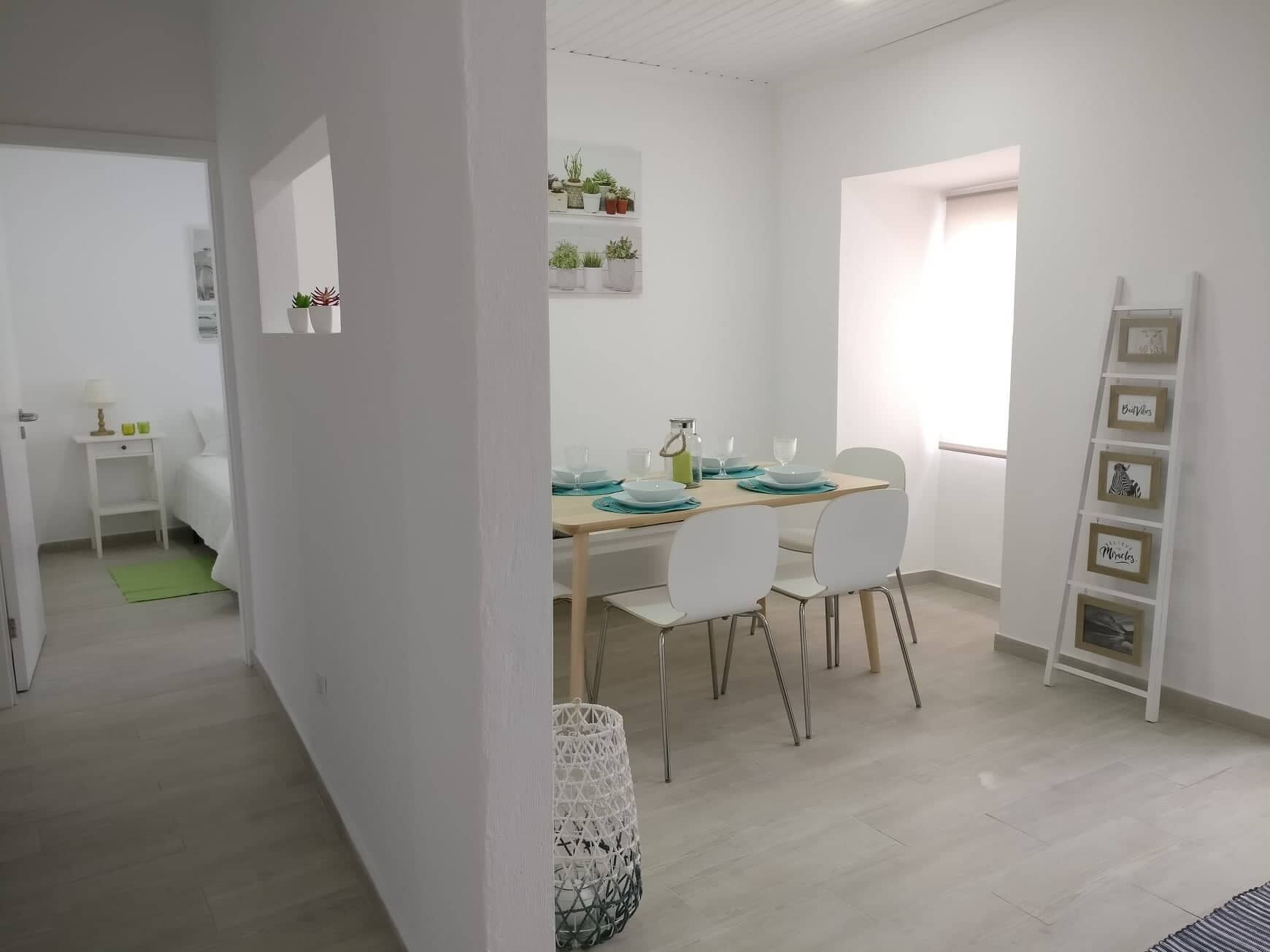 Een moderne eetruimte met een houten tafel voor vier personen, witte stoelen en een bijzettafeltje met een lamp en kopjes.