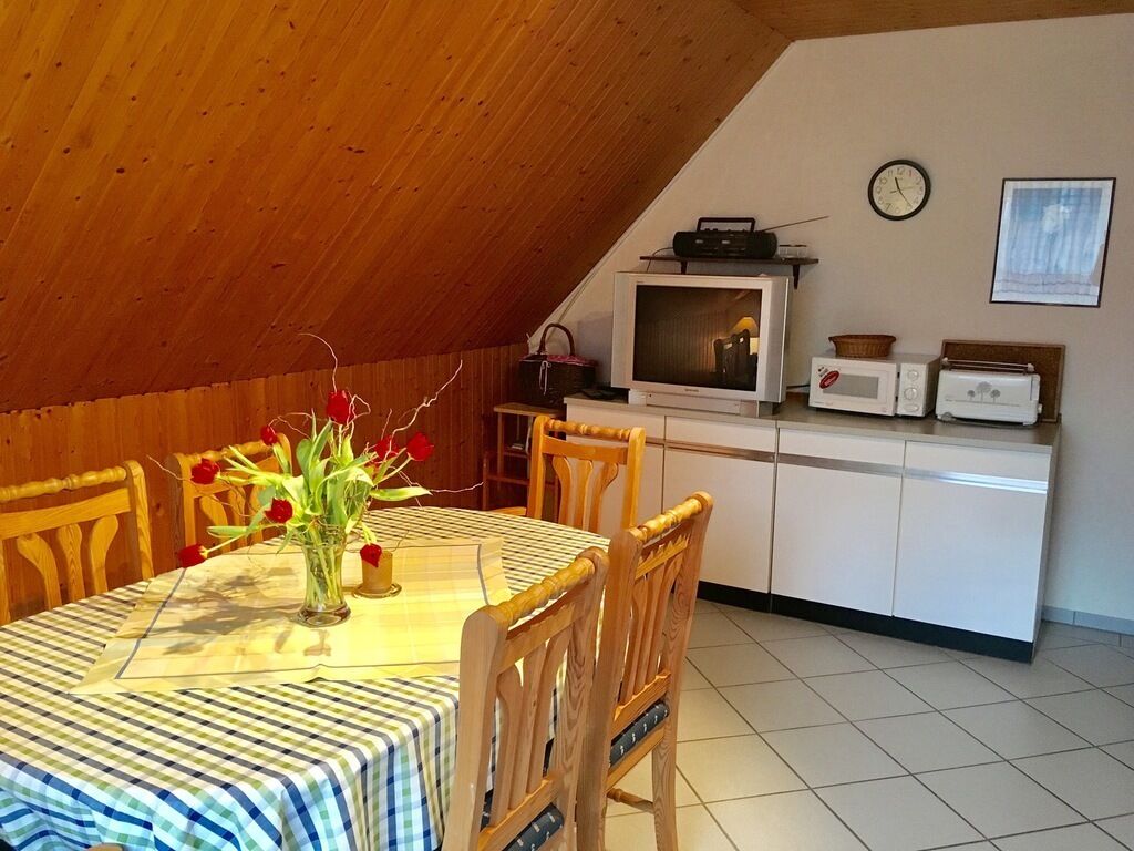 A dining area with a table set for two, wooden chairs, a vase with flowers, and a kitchen area with a microwave, toaster, and a television.