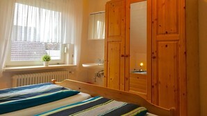 A bedroom with a bed, a wooden wardrobe, a window with curtains, a radiator, and a sink. - Quiet upper apartment in the middle of Norddeich for 4 people + baby (Norden)